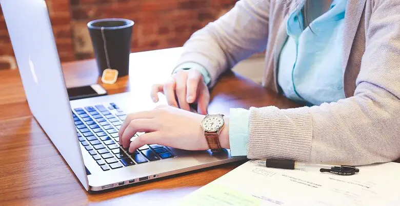 Woman sat at a table working on her laptop with an open notebook and pen next to her