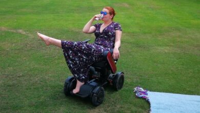 Hannah Barham-Brown in her wheelchair in a park wearing a long black dress with a floral pattern one leg in the air and drinking from a cup