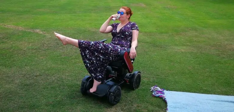 Hannah Barham-Brown in her wheelchair in a park wearing a long black dress with a floral pattern one leg in the air and drinking from a cup