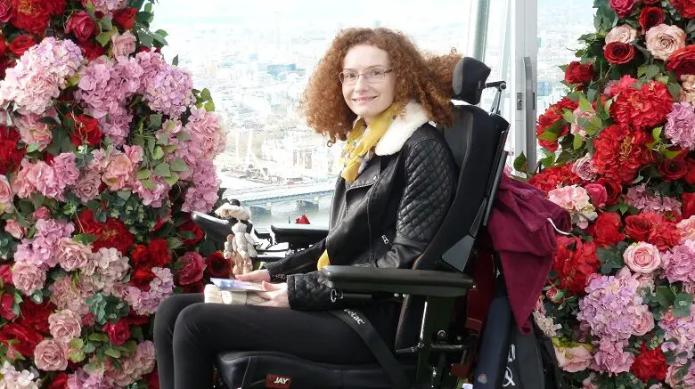 Hannah Deakin with red curly hair in her wheelchair wearing black trousers and a black leather jacket in front of an archway of pink and red flowers