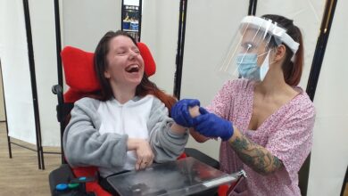 Woman in a wheelchair and grey jumpsuit with a carer wearing plastic gloves, a medical mask and face shield