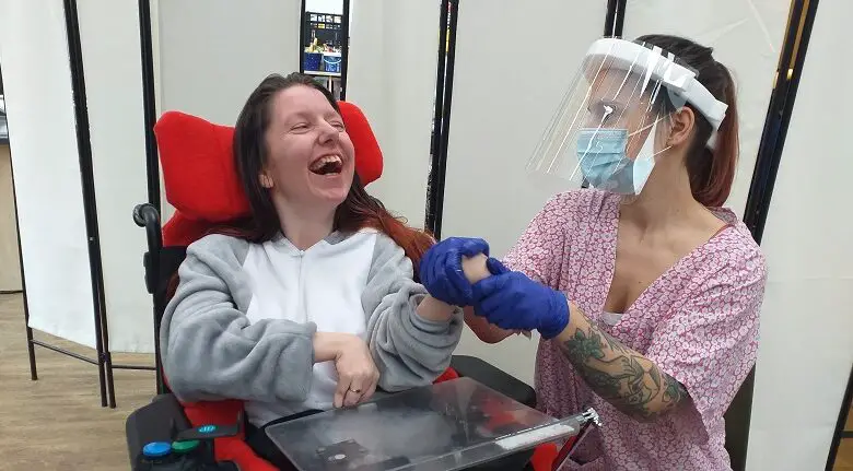 Woman in a wheelchair and grey jumpsuit with a carer wearing plastic gloves, a medical mask and face shield