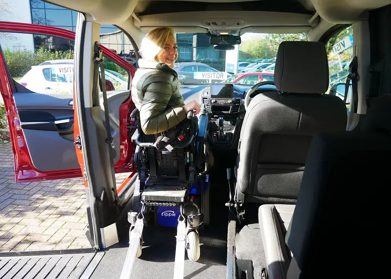 A woman sat in her wheelchair in the passenger seat in a wheelchair accessible vehicle viewed from inside the back