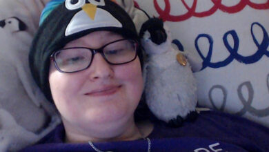 Danni wears a penguin hat and has a toy penguin rested on their shoulder. They are laying in bed.