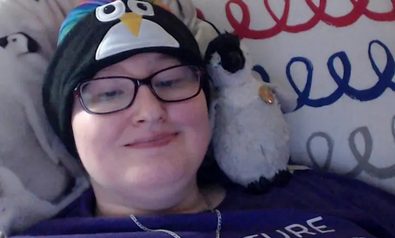 Danni wears a penguin hat and has a toy penguin rested on their shoulder. They are laying in bed.