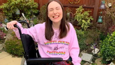 Rebecca is sat in the garden in her wheeelchair, she wears a pink jumper