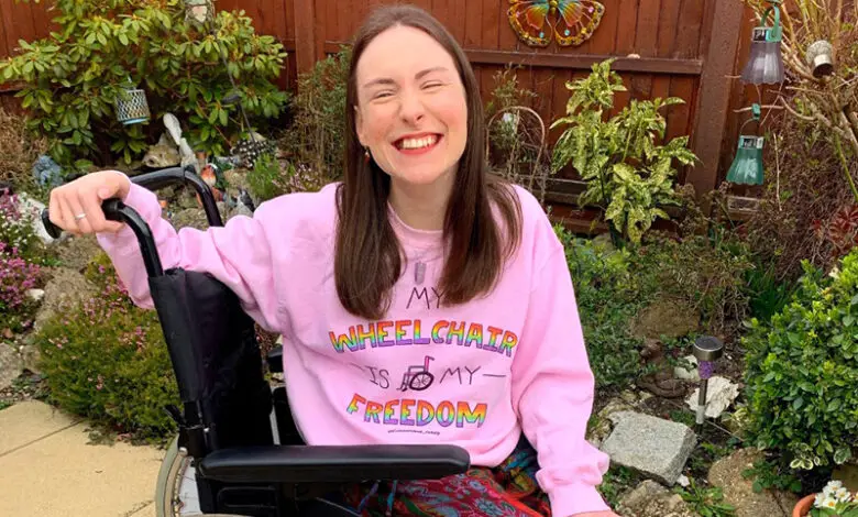 Rebecca is sat in the garden in her wheeelchair, she wears a pink jumper