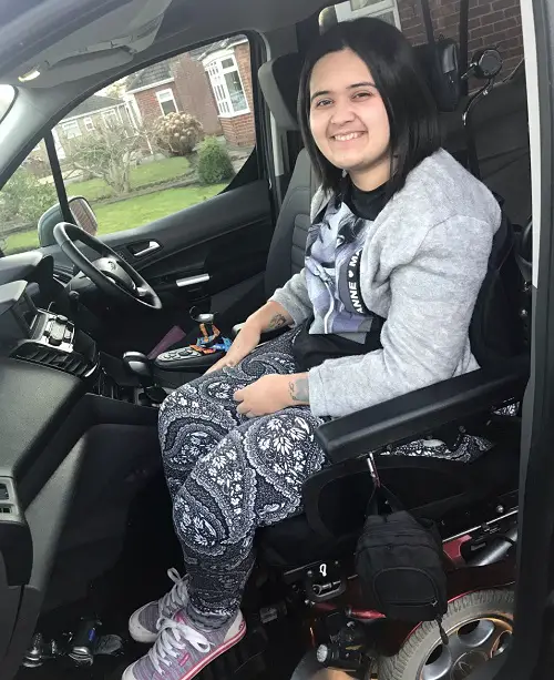 Stefanie Leaf sat in her wheelchair upfront in her wheelchair accessible vehicle wearing back and white leggings, a black t-shirt and grey cardigan