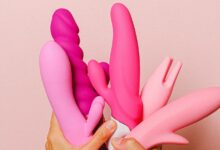 Women's hands holding a collection of pink sex toys and dildos in the air