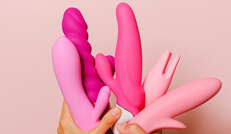 Women's hands holding a collection of pink sex toys and dildos in the air