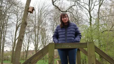 Holly-stood-on-a-wooden-bridge-with-hands-in-pockets-looking-down-she-is-wearing-a-blue-padded-jacket-and-blue-jeans-there-is-a-mass-of-green-trees-in-the-background-clouds-can-be-seen