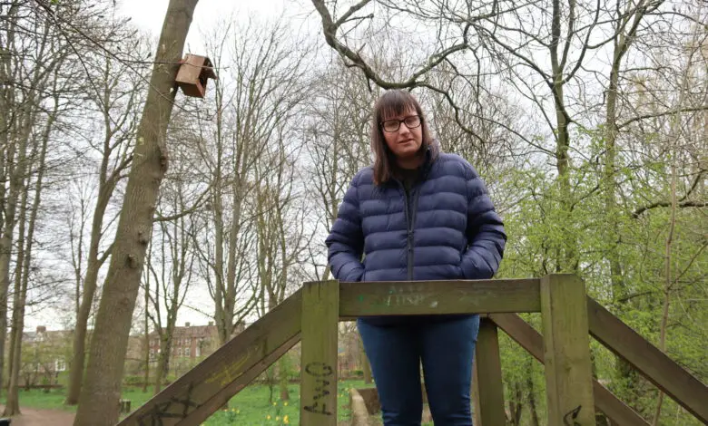 Holly-stood-on-a-wooden-bridge-with-hands-in-pockets-looking-down-she-is-wearing-a-blue-padded-jacket-and-blue-jeans-there-is-a-mass-of-green-trees-in-the-background-clouds-can-be-seen