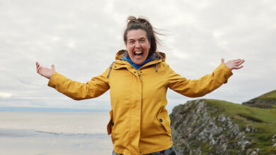 Rosie Jones wearing yellow coat with both arms in the air on Mission Accessible