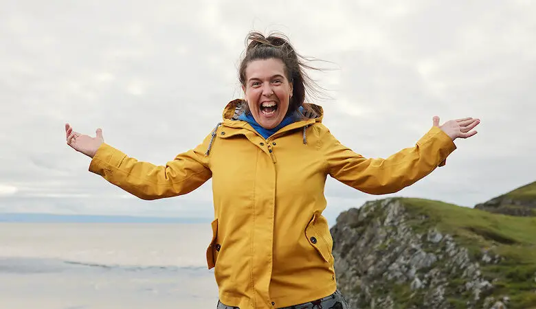 Rosie Jones wearing yellow coat with both arms in the air on Mission Accessible