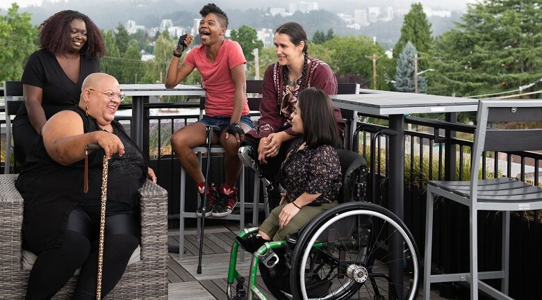 Five women sat around a table outside - one wheelchair user, one using a stick, one visually impaired and one with a proshthetic limb all of different nationalities