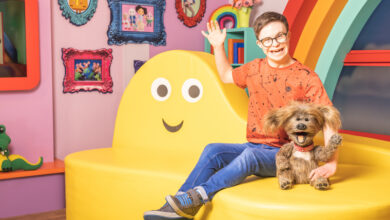 George Webster in CBeebies studio wearing orange T-shirt, blue jeans and glasses