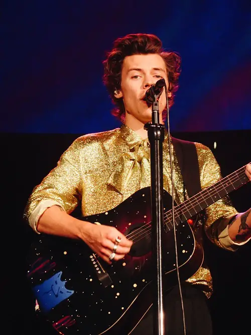 Harry styles in a gold shirt stood at a microphone singing on stage