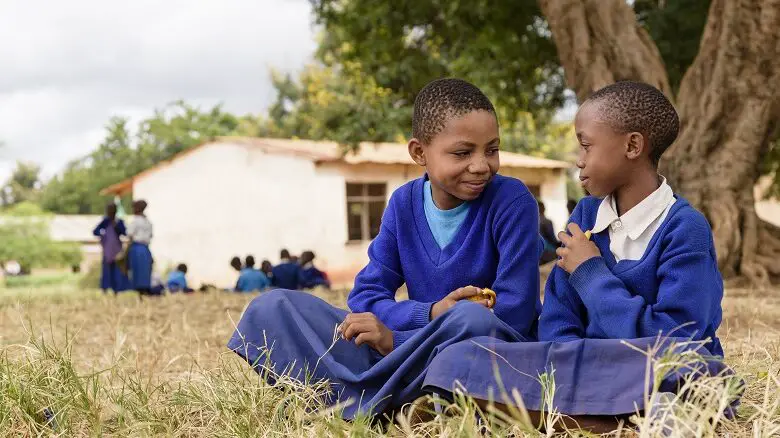 Two young girls wearing school uniforms sitting on the floor outside a school in Africa