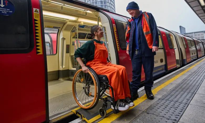 Photograph of a young woman in bright orange dungarees and a green long sleeve top, alighting a London train using a bridging device, whilst being greeted by a smiling member of transport staff.