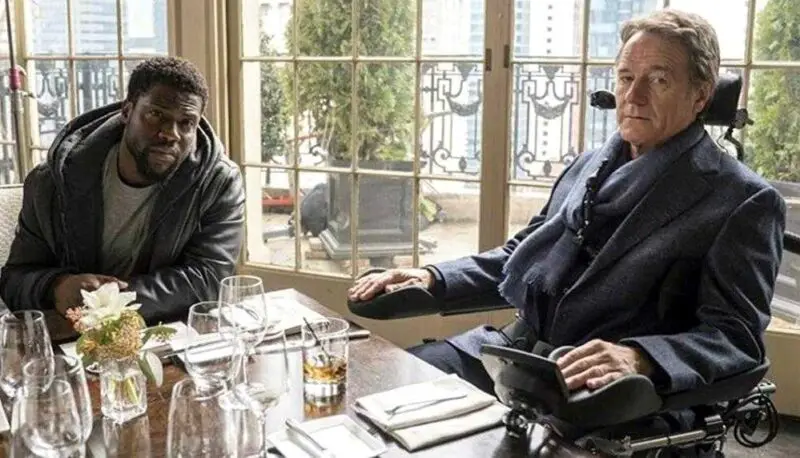 Bryan Cranston playing wheelchair user Phillip Lacasse sat in a restaurant with Dell played by Kevin Hart in the film Upside Down on Amazon Prime Video