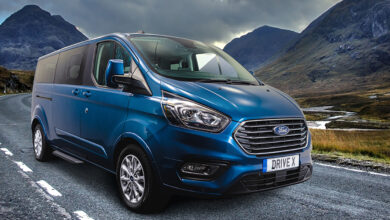 Blue Ford Drive X wheelchair accessible vehicle from Sirus driving along a road with mountains in the background