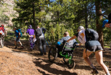 A group of people, including a wheelchair user in a Mountain Trike all-terrain wheelchair, hiking through a forest