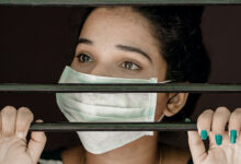 A woman with a face mask on looking through bars with her hands holding them