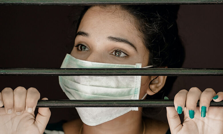 A woman with a face mask on looking through bars with her hands holding them