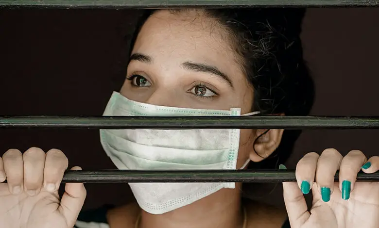 A woman with a face mask on looking through bars with her hands holding them