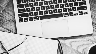 Black and white image showing a laptop keyboard, writing pad, pen and cup of coffee from above
