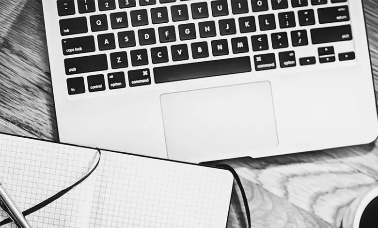 Black and white image showing a laptop keyboard, writing pad, pen and cup of coffee from above