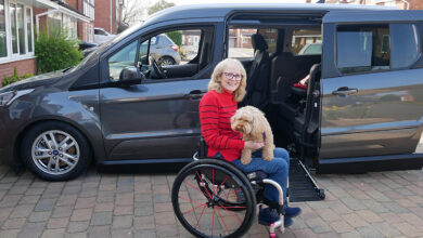 Wheelchair user Susan wearing a red and black top with her dog on her lap on her driveway in front of her Sirus Ford Grand Tourneo Connect wheelchair accessible vehicle