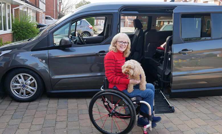 Wheelchair user Susan wearing a red and black top with her dog on her lap on her driveway in front of her Sirus Ford Grand Tourneo Connect wheelchair accessible vehicle