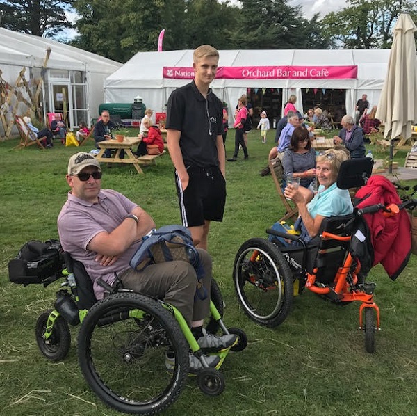 A man and a woman in all-terrain Mountain Trike wheelchairs at an outdoor festival