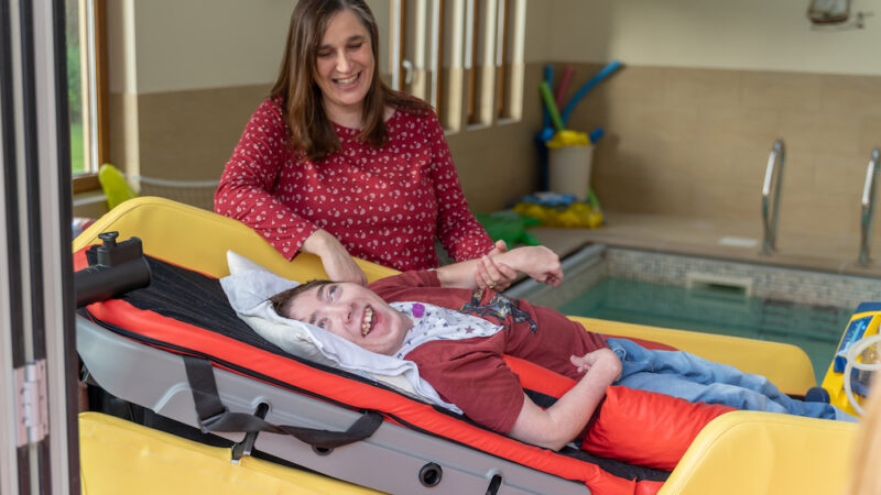 Skiggle Co-founder Christine with long brown hair wearing a red top with her son James lying on an adjustable bed in front of a hydrotherapy pool smiling