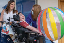 Skiggle Co-founders Christine and Helen with James who is sat in his wheelchair smiling with a large beach ball on his lap