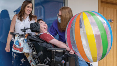 Skiggle Co-founders Christine and Helen with James who is sat in his wheelchair smiling with a large beach ball on his lap