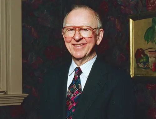 Edwin Krebs wearing large glasses, a black jacket and patterned blue and red tie standing in front of a patterned wallpaper 