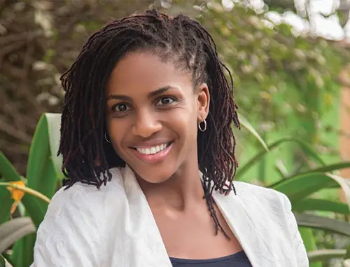 A close up of Farida Bedwei with black hair and small hoop earrings wearing a white jacket and a black top stood outside in front of a tree