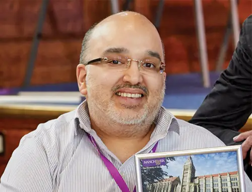 A close up of Hamied Haroon with short grey hair and facial stubble wearing glasses and a pale blue shirt holding a book and sat in a lecture theatre