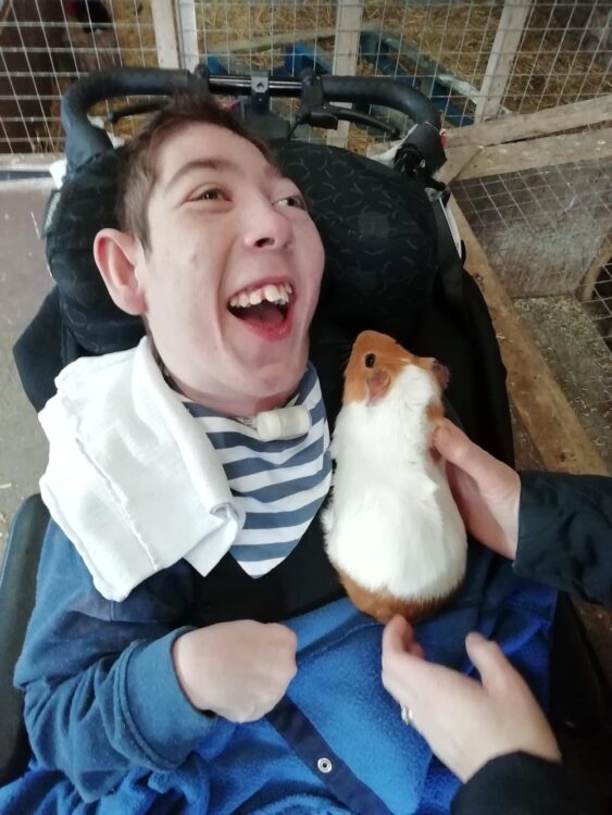 James Singleton smiling in his wheelchair wearing a blue top with a white and ginger guinea pig on his lap 
