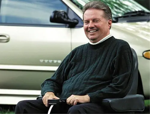 Ralph Braun in a wheelchair wearing a black jumper with short brown hair in front of a car