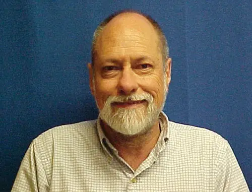A close up of Richard Mankin with short grey hair and a short grey beard wearing a white shirt stood in front of a blue background