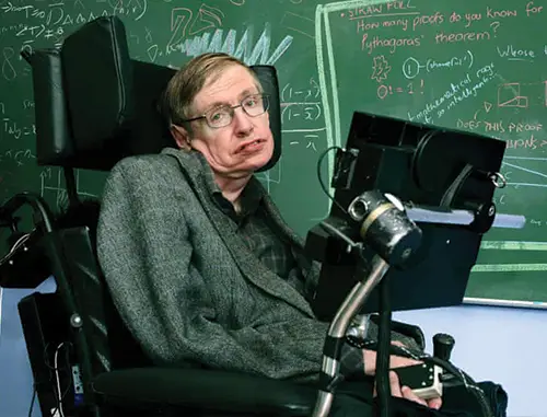 Stephen Hawking in his wheelchair with short brown hair wearing a grey suit jacket and glasses in front of a blackboard with equations on it in chalk