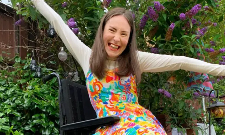 Rebecca with brown hair wear a colourful dress with a with polo, red shoes and a colourful beaded necklace. She sits in her wheelchair, with rainbow spoke guards, with her arms raised above her head. Rebecca is in the garden surrounded by flowers.