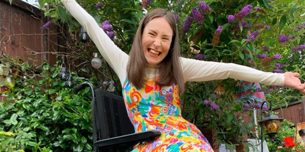 Rebecca with brown hair wear a colourful dress with a with polo, red shoes and a colourful beaded necklace. She sits in her wheelchair, with rainbow spoke guards, with her arms raised above her head. Rebecca is in the garden surrounded by flowers.
