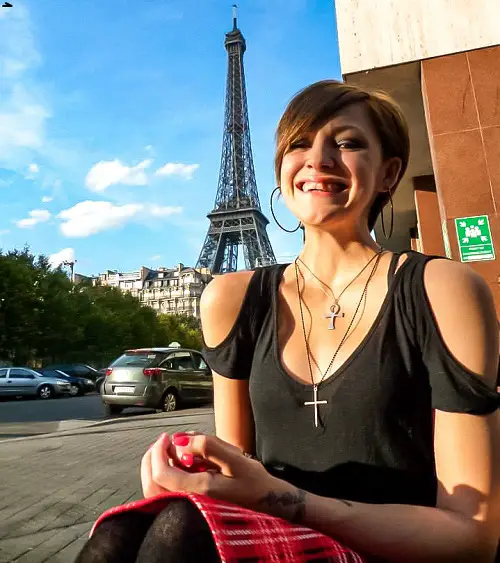 Molly Gudger with short brown hair, wearing a black top that's off the shoulders, a red tartan skirt and large hoop earrings and two necklaces, in her wheelchair in Paris at the side of a street with the Eiffel Tower in the background in the distance