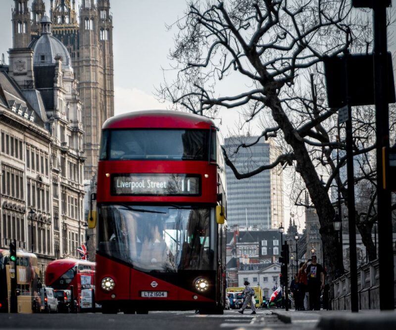 A London bus viewed from the front driving down a street in London with buildings on the left and trees on the right