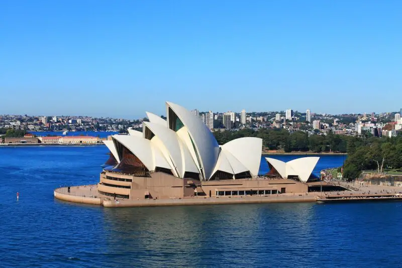 The Sydney Opera House surrounded by water with city buildings in the background. The building is white with sculptural domes