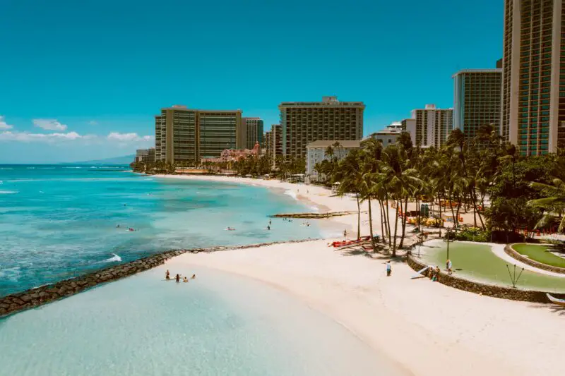 Waikiki beach in Honolulu, Hawaii with crystal blue water, white sand and tall buildings on the water's edge with palm trees dotted along the beach